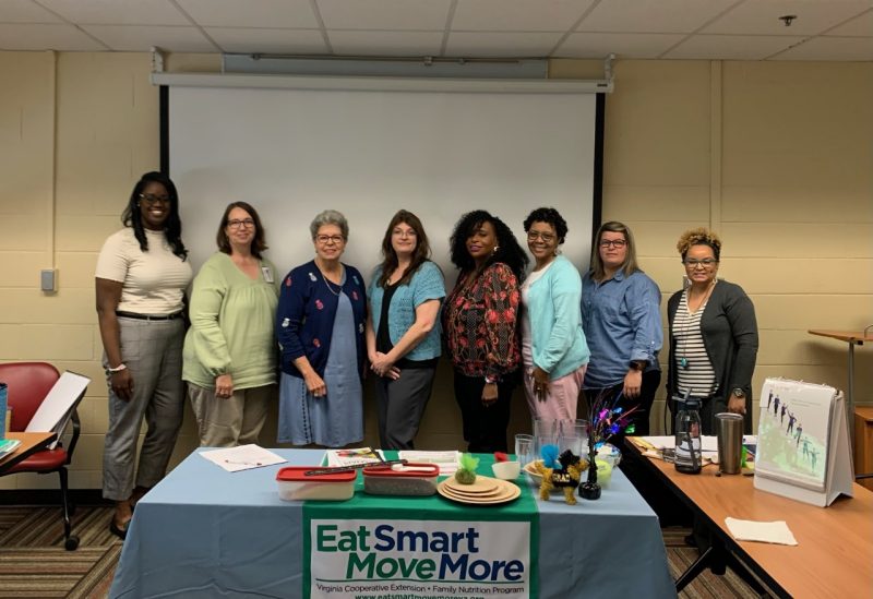 A group of eight extension faculty and staff stand inside of a classroom behind a table to smile for a photo.