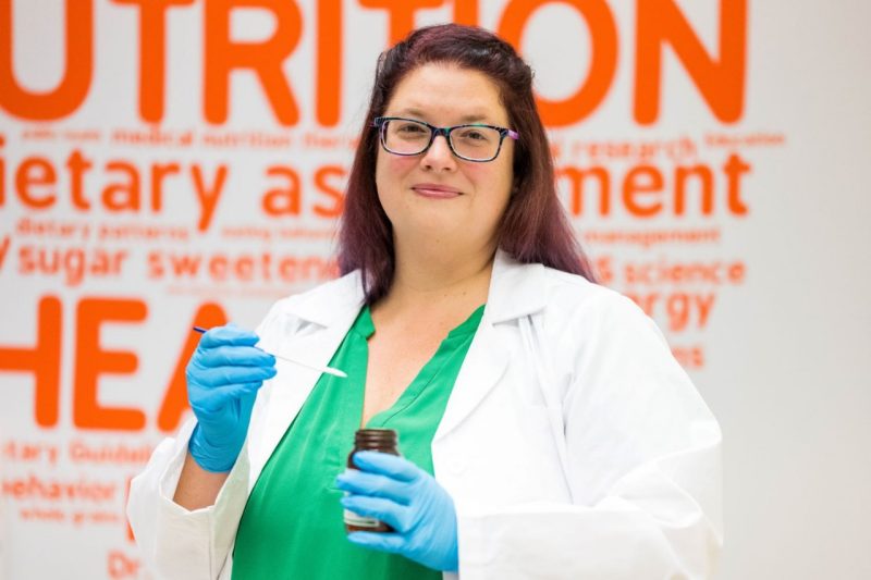 A professor stands with a lab coat on measuring a spoonful of sugar.