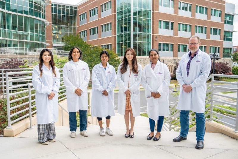 A group of six researchers wear lab coats and stand in a line at Virginia Tech's Carillion campus.