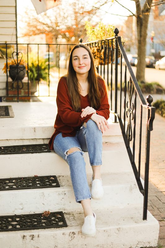 A Virginia Tech HNFE Ambassadors sits on the top of the stairs outside on her porch and smiles for a photo learning against the handrail.